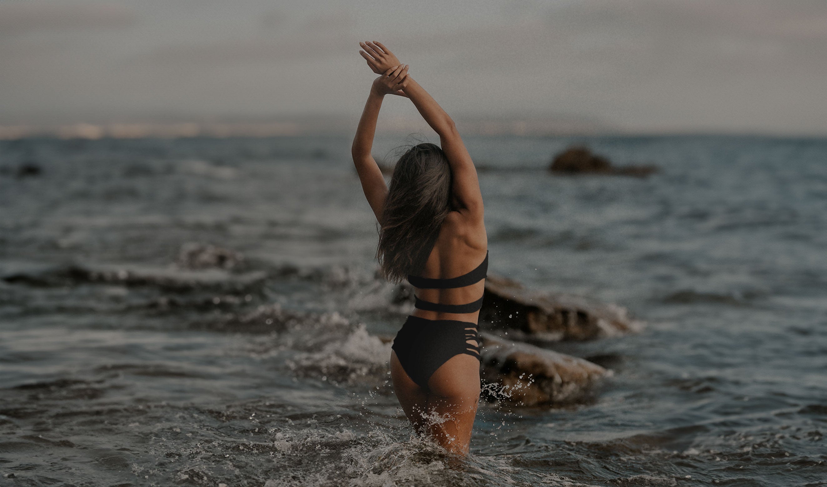 Model wearing Rhea Bottom in black swimsuit standing in the ocean with arms raised.