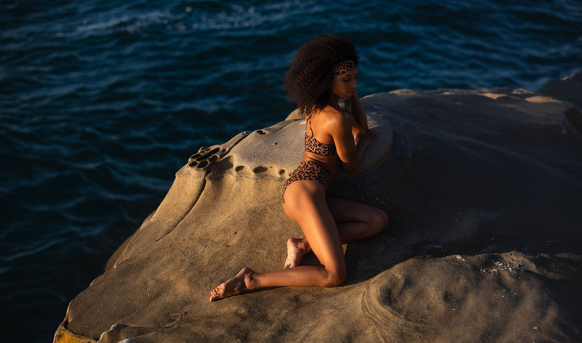 A model wearing the Leopard Sol print Lila Top and Mia Bottom, kneeling on coastal rocks in the warm afternoon sunlight.